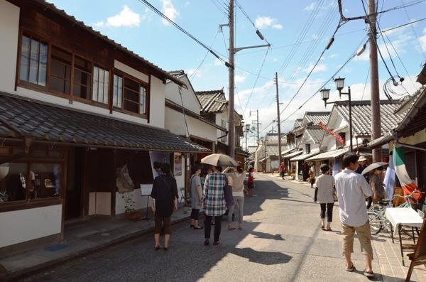 日本家屋が右側にも左側にも並んでいて、中央に通っている道を10名ほどの人達が歩いている写真